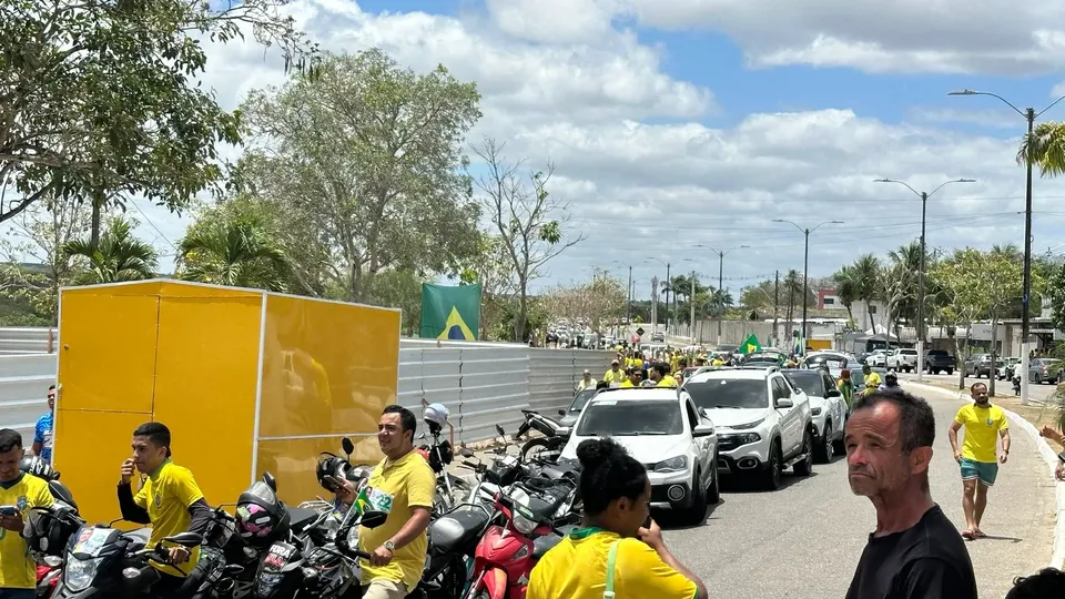 Fila de carros e motos na avenida da Pista Nova, em Macaíba, com apoiadores de camisa amarela e uma bandeira do Brasil ao fundo