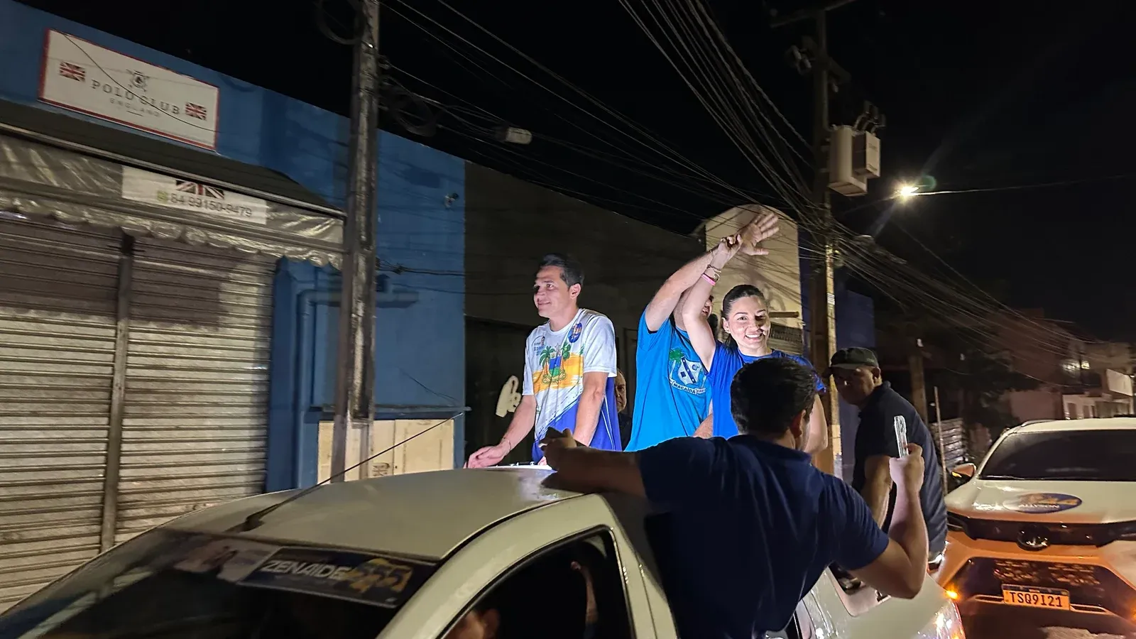 Três pessoas acenam em cima de uma caminhonete branca durante carreata noturna em rua de Macaíba