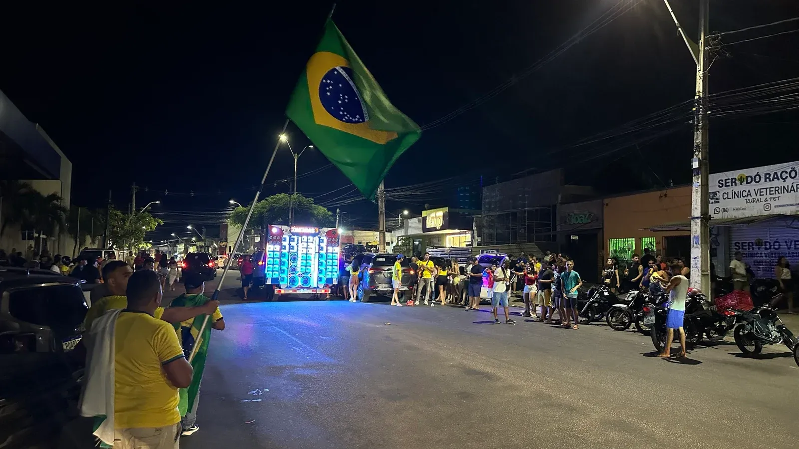 Apoiador agita bandeira do Brasil em rua do Centro de Macaíba, à noite, com paredão de som iluminado e pessoas de verde e amarelo