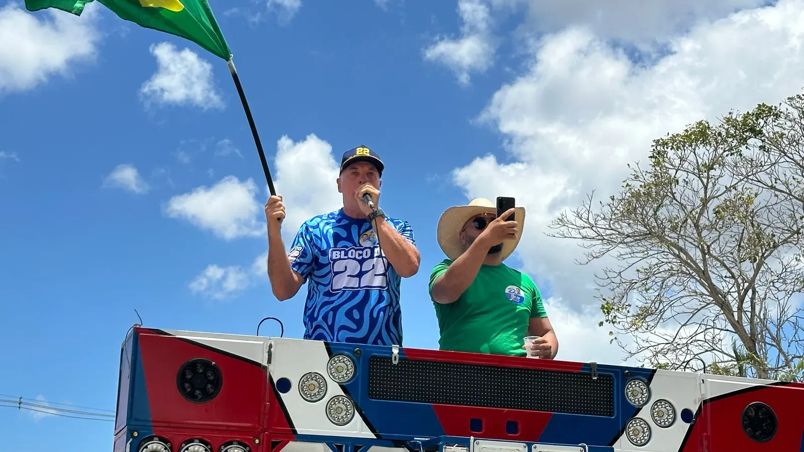 Coronel Hélio, de boné e camisa azul com o número 22, discursa ao microfone em cima de um trio e segura uma bandeira do Brasil
