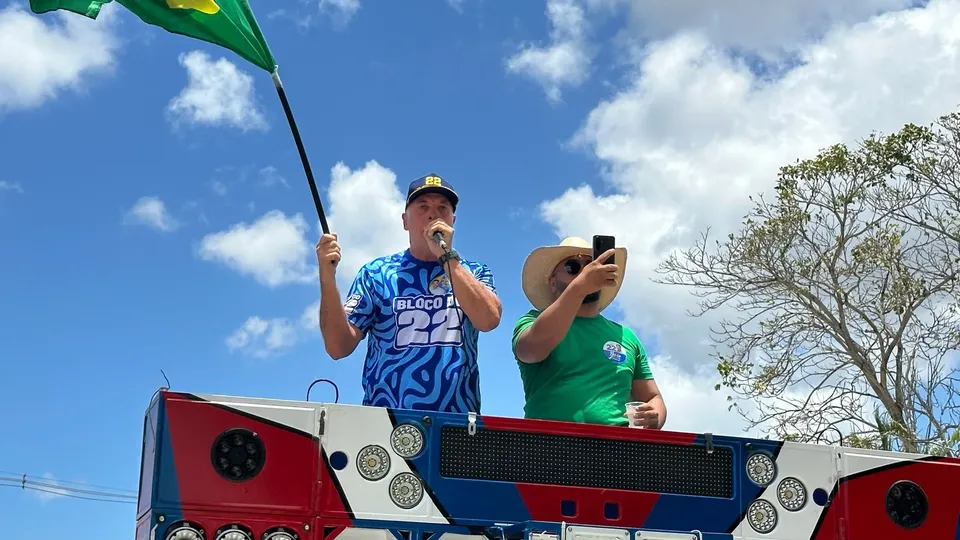 Coronel Hélio, de boné e camisa azul com o número 22, discursa ao microfone em cima de um trio e segura uma bandeira do Brasil