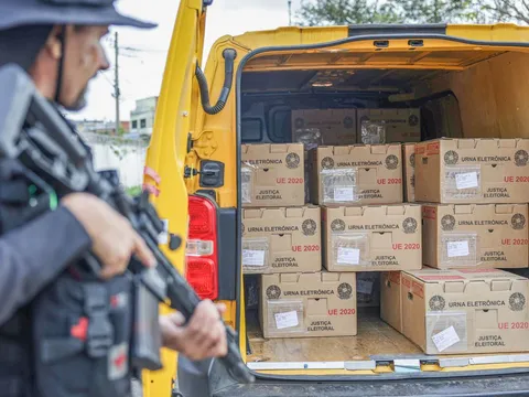 Caixas de urnas eletrônicas em uma van, com um policial ao lado