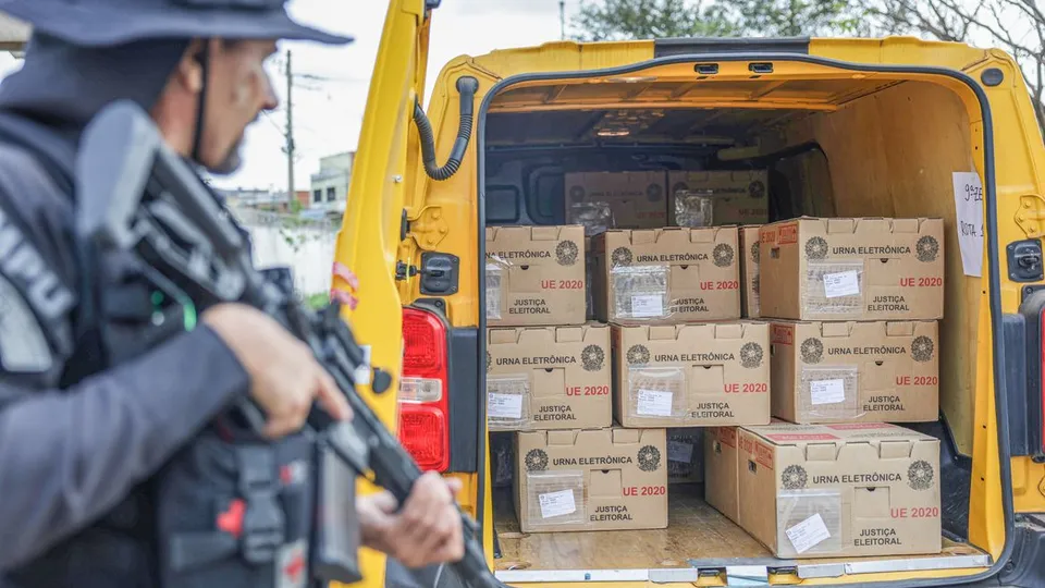 Caixas de urnas eletrônicas em uma van, com um policial ao lado