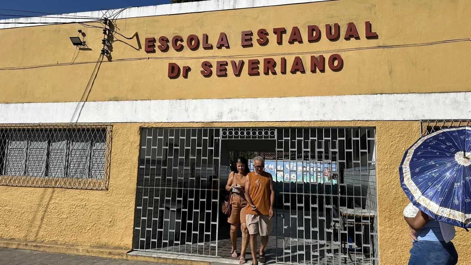 Casal sai pelo portão gradeado da Escola Estadual Dr. Severiano, de fachada amarela, enquanto uma mulher passa com guarda-chuva