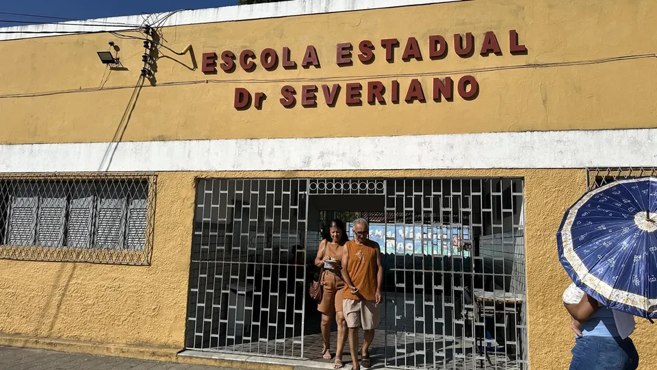 Casal sai pelo portão gradeado da Escola Estadual Dr. Severiano, de fachada amarela, enquanto uma mulher passa com guarda-chuva