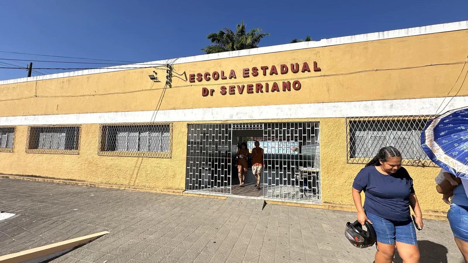 Fachada amarela da Escola Estadual Dr. Severiano com portão aberto e duas pessoas saindo; uma mulher passa na calçada com capacete na mão