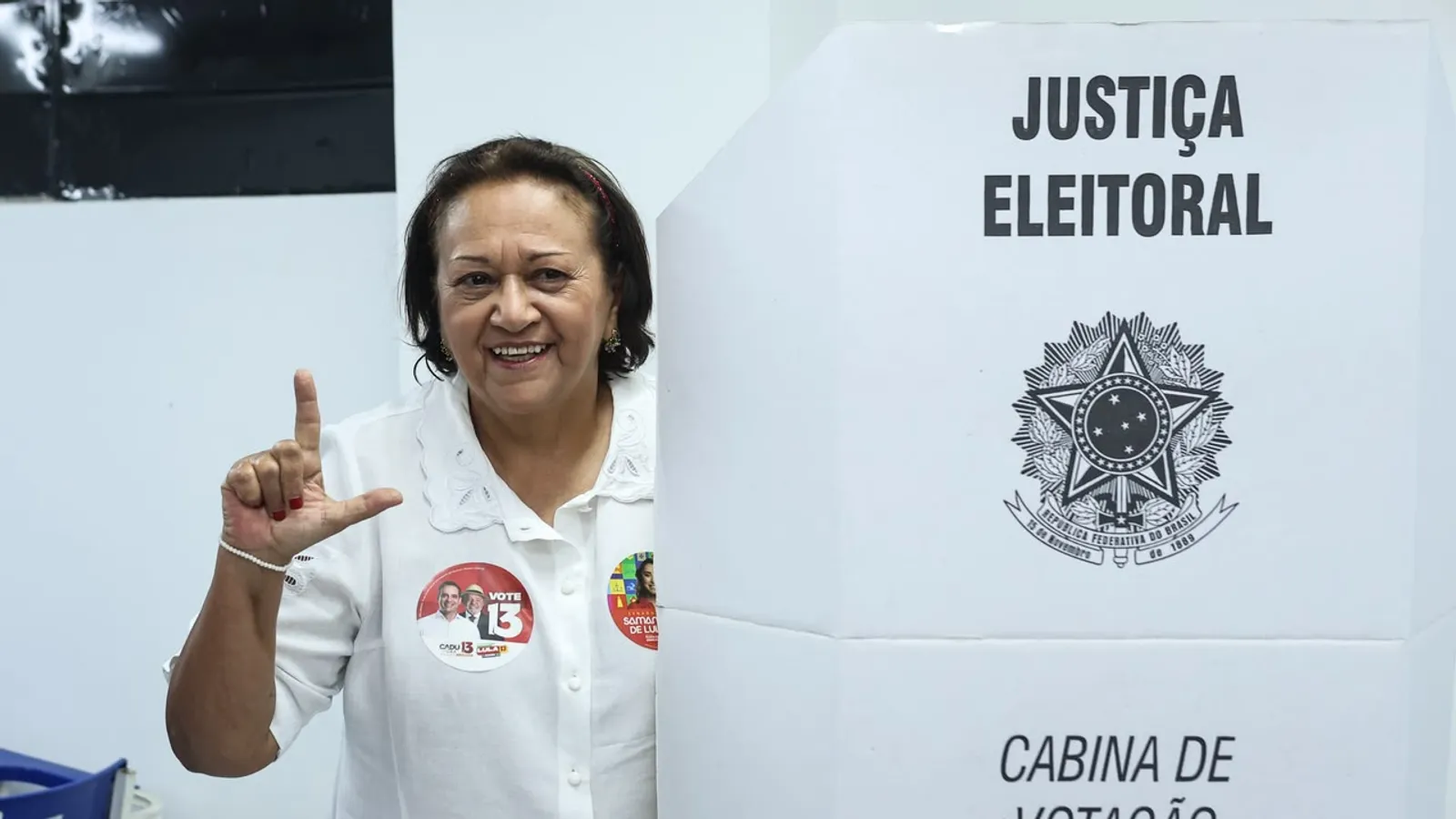 Fátima Bezerra sorri e faz o gesto do L ao lado da cabine de votação