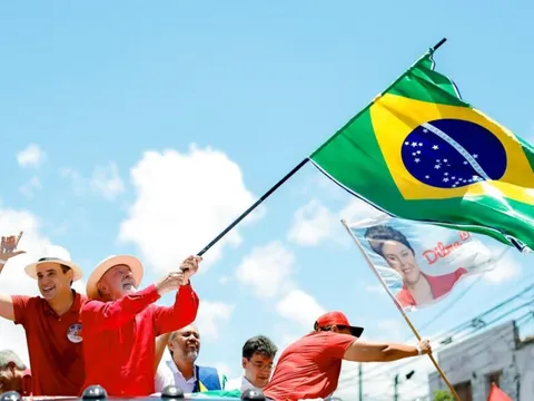 Lula, de camisa vermelha e chapéu, agita bandeira do Brasil em carro aberto ao lado de Cadu, no Alecrim