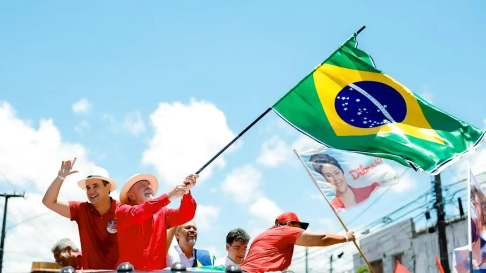 Lula, de camisa vermelha e chapéu, agita bandeira do Brasil em carro aberto ao lado de Cadu, no Alecrim