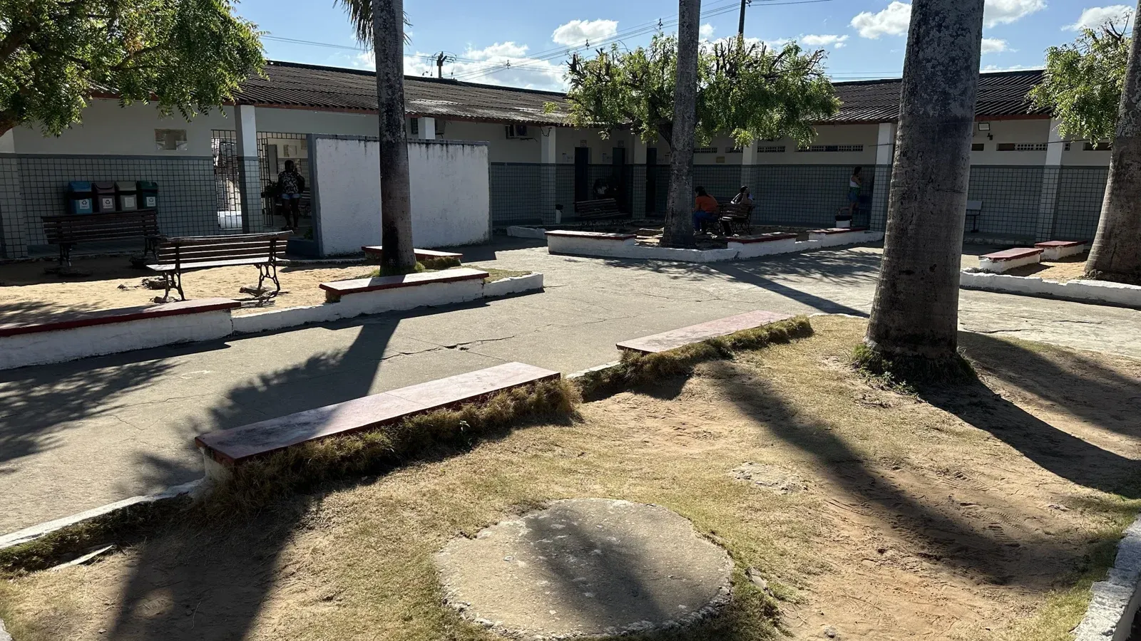 Pátio arborizado da escola com bancos de alvenaria e poucas pessoas sentadas na sombra, junto às salas