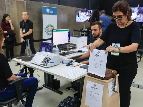 Servidora da Justiça Eleitoral segura cédula ao lado de urna eletrônica durante teste de integridade